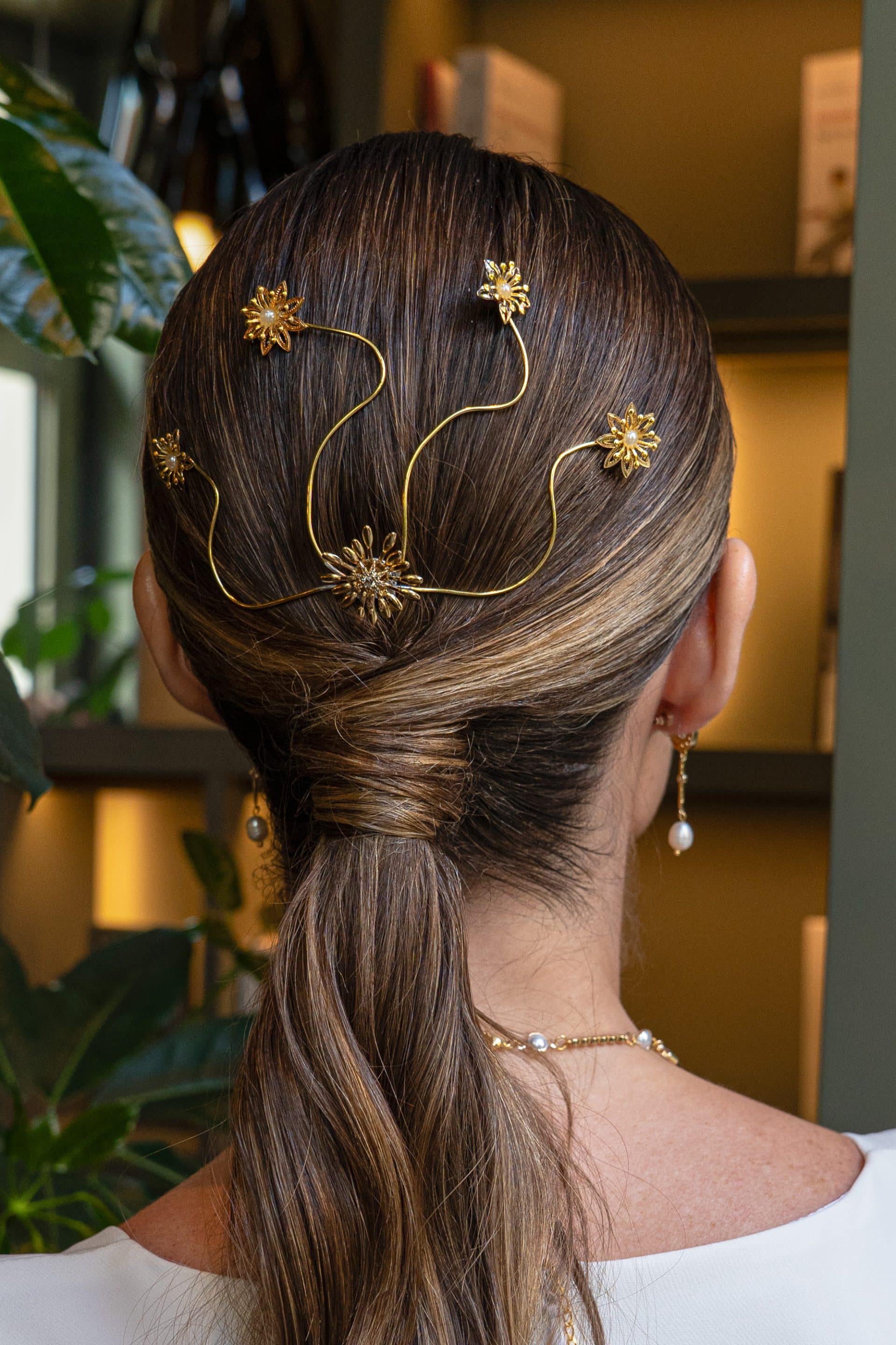 Vista trasera del peinado de una modelo con coleta baja decorada con el tocado dorado flexible Judith de flores y perlas moldeables.